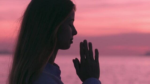 Calm woman stands praying at beach during pink sunset on sea. Female prayer in religion. Concept of true Faith in God. Religion, spiritual and hope concept. Pray to God.