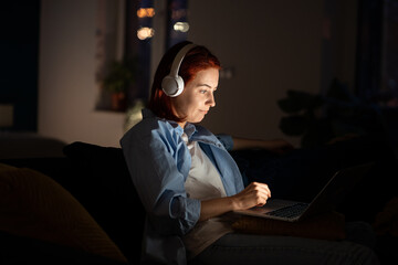 Focused freelancer woman in headphones sitting on sofa, working on laptop at home late night. Side...