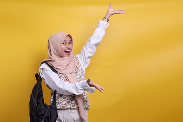Excited Asian muslim student girl in hijab carrying backpack showing empty space for text or ads with both hands