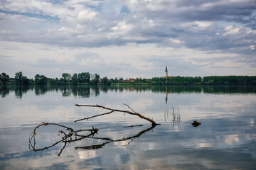 Rural view of the old cathedral tower with reflection in the water and cottages against the background of a wide river and cloudy sky. Balkan view of the Danube