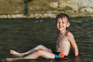 a preschool-age boy in bathing trunks plays sitting in the river