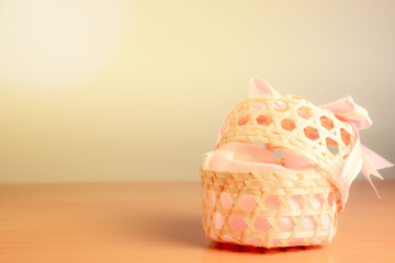 A woven wooden basket with an open lid and a pink bow-tie placed on the right side of the warm, white background