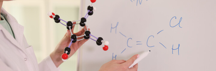 Female chemist holding molecular model and drawing chemical formulas on whiteboard.