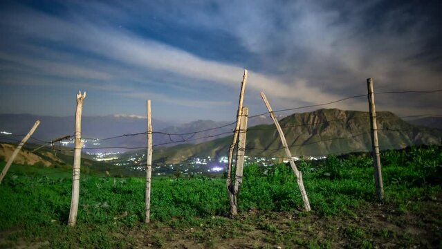 Night in mountains timelapse. Night sky with moving clouds taken on top of mountain covered with greenery. Old rickety fence. Blue night sky, lights of residential settlement. Time for dreams