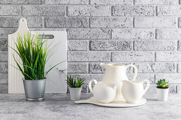 Beautiful stylish kitchen background in light colors. A set of white jugs, cutting boards and houseplants on a gray stone countertop. Gray brick wall.