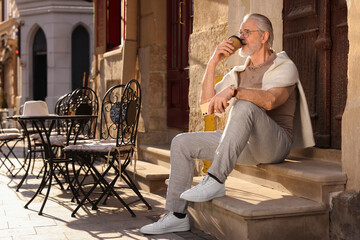 Handsome senior man sitting on doorstep with smartphone and drinking coffee outdoors, space for text