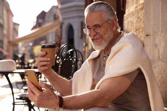 Handsome senior man sitting on doorstep, using smartphone and drinking coffee outdoors, space for text