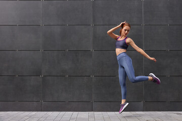 Beautiful woman in gym clothes jumping near dark grey wall on street, space for text