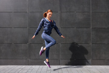 Beautiful woman in gym clothes jumping near dark grey wall on street, space for text