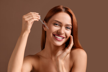 Fototapeta premium Beautiful young woman applying cosmetic serum onto her face on brown background