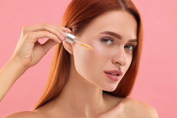 Fototapeta premium Beautiful young woman applying cosmetic serum onto her face on pink background