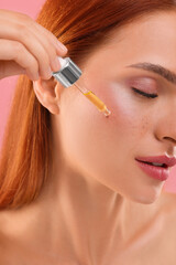 Fototapeta premium Beautiful young woman applying cosmetic serum onto her face on pink background, closeup