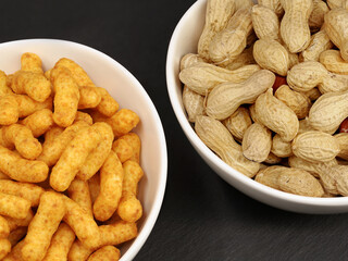 Portion of peanut flips and naturally roasted inshell peanuts in white bowls on black background, snack comparison of peanuts