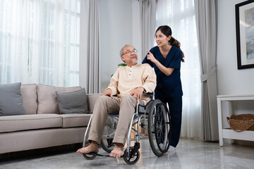 Fototapeta na wymiar A young Asian female nurse or physical therapist in a blue uniform, standing and talking happily behind a senior Asian man sitting on a wheelchair while pushing him to relax in the house living room.