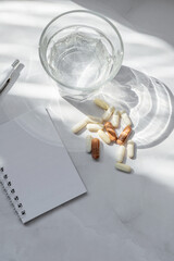 Water glass, pills and capsules, pen and blank notebook mock up on white table background with aesthetic sunlight shadows. Health care, wellness concept, diet planning