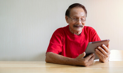 80s grey haired old man sit at wooden table hold tablet and looking at screen makes call to physician