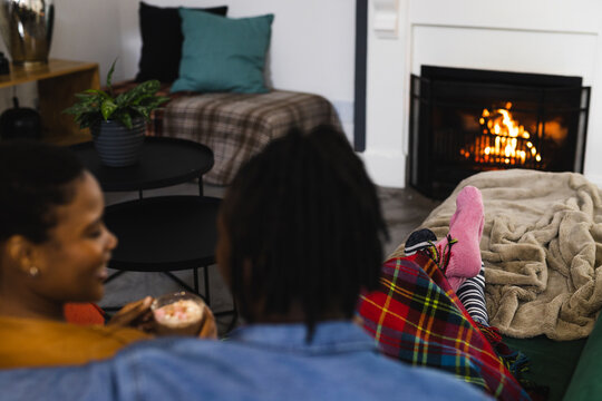 Happy african american couple in socks relaxing at home in front of open fire with hot chocolate