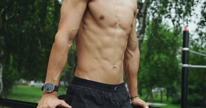 Cropped view on athletic man training on parallel bar and doing abs exercise outdoors at sport ground. Closeup on male with shirtless torso and perfect abdominal muscles at workout.