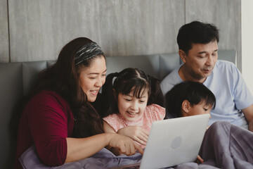 Fototapeta premium Happy Asian family lying on bed in bedroom with smile, father and mother with daughter and son, lovely parents using laptop computer for education and spent quality time together, laughing together