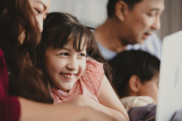 Happy Asian family lying on bed in bedroom with smile, father and mother with daughter and son, lovely parents using laptop computer for education and spent quality time together, laughing together