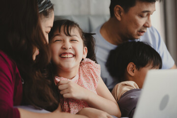 Happy Asian family lying on bed in bedroom with smile, father and mother with daughter and son, lovely parents using laptop computer for education and spent quality time together, laughing together