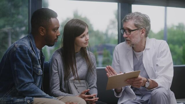 Multicultural couple sits on couch in clinic lobby area after appointment with doctor. Doctor talks with patients about medical test results. Waiting room in modern medical center. Healthcare system.
