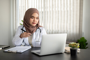 A beautiful Asian Muslim female doctor is working on her laptop at her desk at a hospital or clinic.
