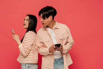 Asian man looking at his girlfriend's mobile phone screen while standing isolated over red background