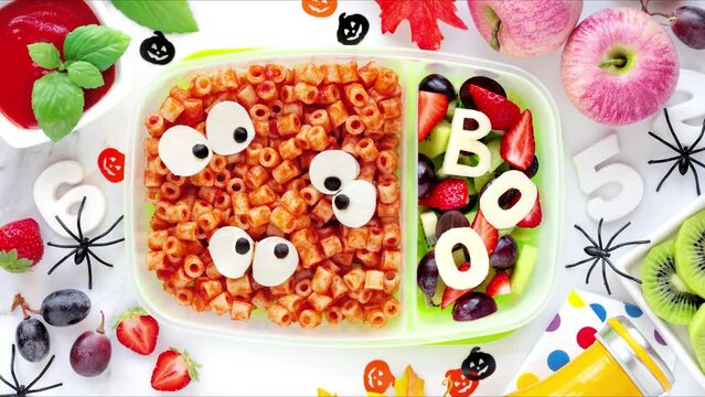 School lunch box for Halloween. Fun food for kids - pasta with tomato sauce with spooky ghost eyes and fruit salad. Stop motion animation