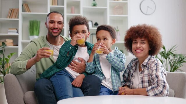 Adorable multiracial kids in casual wear drinking fresh juice while mom and dad sitting aside in cozy interior. Cheerful young family of four planning after dinner walk in nearby park on summer day.