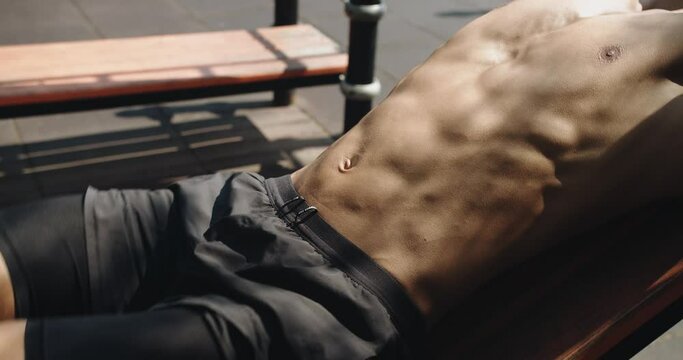 Closeup on shirtless man with athletic body doing reverse crunch exercise for abs muscles on bench at outdoors gym. Cropped view on young male with six pack has workout and abdominal training
