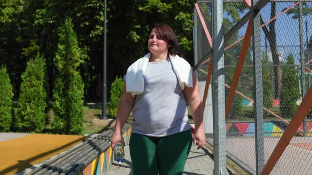 Happiness fills the air as an overweight woman takes a post workout break outdoors. She smiles with satisfaction, holding a water bottle and a towel around her neck.