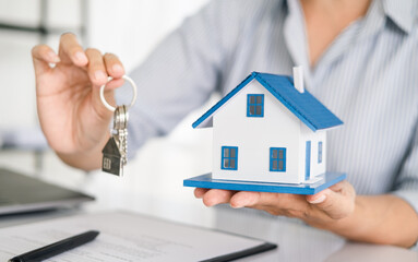 Real estate agent holding house key on house shaped keyring on table with house designs document, calculator, model house. Concept of Investment property.