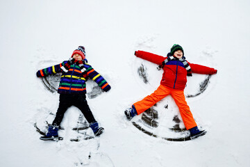 Two little siblings kid boys in colorful winter clothes making snow angel, laying down on snow. Active outdoors leisure with children in winter. Happy brothers with warm hat, gloves, winter fashion.