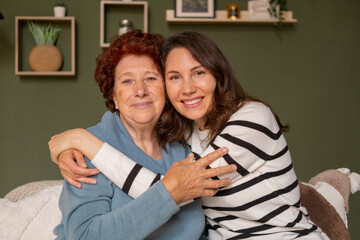 Portrait of a cheerful grandmother and granddaughter sitting on the sofa during the day in the living room and hugging each other tightly with their arms