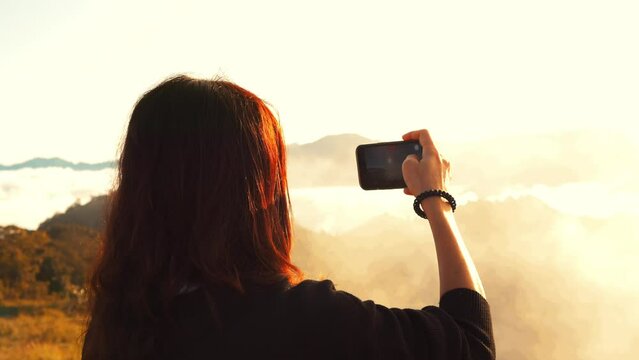 Young girl standing on the top of a mountain and making photos and videos of beautiful sunrise over morning clouds using a mobile phone at Phu Chi Fa viewpoint, Thailand.