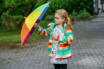 Little girl on way to elementary school or kindergarden. Preschool Child with colorful rainbow umbrella and waterproof jacket with school bag. Kid walking in autumn shower. Outdoor fun by any weather