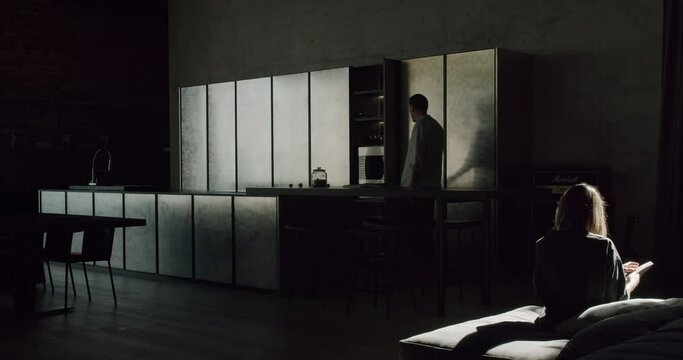 Couple lifestyle. A woman sitting on sofa and reading a book, man entering dark room and close kitchen cupboard in modern apartment with minimalist style.