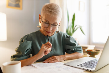 Elderly businesswoman in glasses examining contract of agreement with partners before signing it, looking at paper with concentrated face touching chin with pen sitting at cabinet in front of laptop