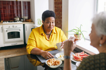 African american senior plus size female sitting at kitchen table in front of her caucasian friend...