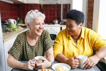 Fototapeta na wymiar Two senior female friends of diverse ethnicity laughing together at funny situation in past or jokes drinking coffee with sweats at kitchen table, recollecting best memories of their lives