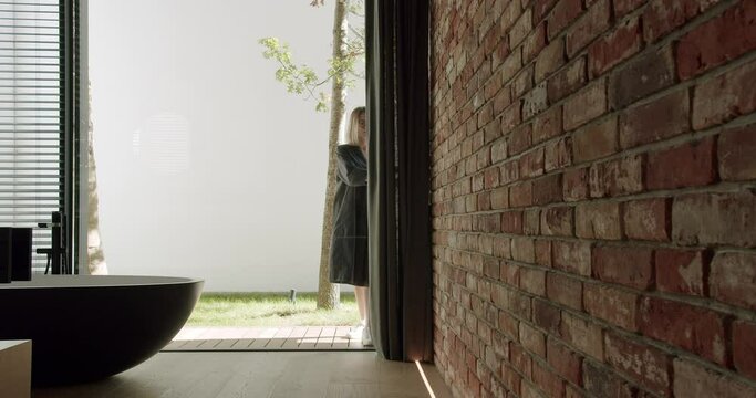 Woman open sliding glass door and enter in room with freestanding bath tub, brick wall and modern loft interior at cottage house.