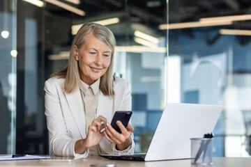 Senior mature experienced woman at workplace inside office, business woman happy smiling using phone sitting with laptop, gray haired woman boss using app on smartphone typing message.