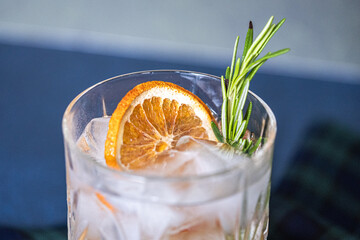 Glass of gin and tonic cocktails with ice, sprig of rosemary and sliced dry orange on a blue...
