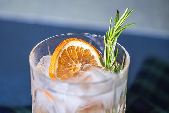 Glass of gin and tonic cocktails with ice, sprig of rosemary and sliced dry orange on a blue background. Copy space