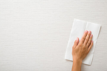 Young adult woman hand holding white dry paper napkin and wiping light gray wallpaper surface. Closeup. Front view. Empty place for text.