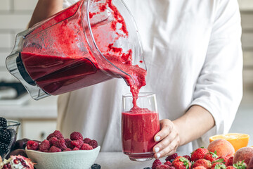 A woman pours a berry smoothie from a blender into a glass.