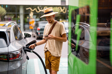Man in hat plugs a cable in electric vehicle, while standing with phone on a public charging station outdoors. Concept of travel by electric car and green energy for driving