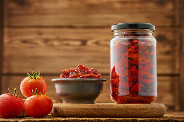 Glass jar and bowl of sun-dried tomatoes in olive oil and fresh tomatoes on a wooden rustic background. Copy space. Studio shot from a low angle.
