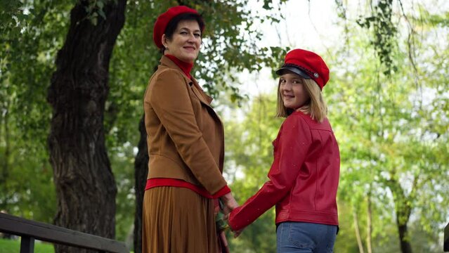 Confident smiling teenage girl and senior woman holding hands looking at camera standing in autumn park. Medium shot portrait of charming grandmother and pretty granddaughter posing outdoors
